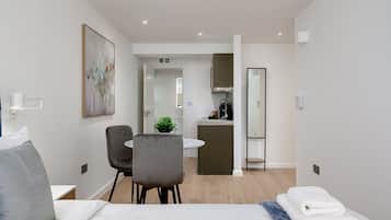 A modern hotel room with a bed, a desk, a chair, a small kitchen area, and a bathroom.