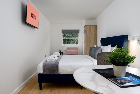 Una habitación de hotel moderna con cama, mesita de noche, planta en maceta y televisor montado en la pared.