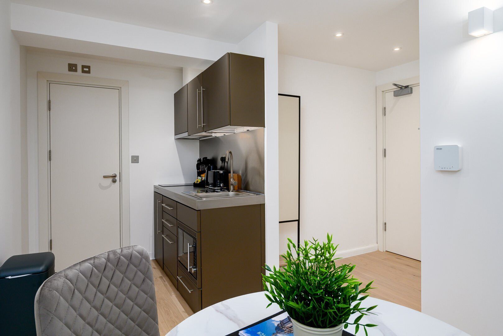 A compact kitchen with a built-in stove, a coffee machine, and a small countertop. There is a chair and a table with a potted plant.