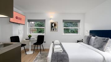 A modern hotel room with a bed, bedside tables, a desk, and two windows.