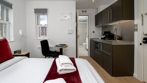 Ett hotellrum med en säng, ett skrivbord, en stol, ett litet bord, ett litet kök och ett badrum som syns genom en öppen dörr.
