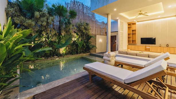 A modern outdoor pool area with lounge chairs, a wooden deck, and a well-lit interior space with a TV and seating.