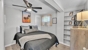 Quarto com cama, ventoinha no teto, janela com cortinas, parede com quadro e estante com fotografias emolduradas.