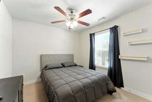 Un dormitorio con cama, ventilador de techo, ventana con cortinas y estanterías empotradas.