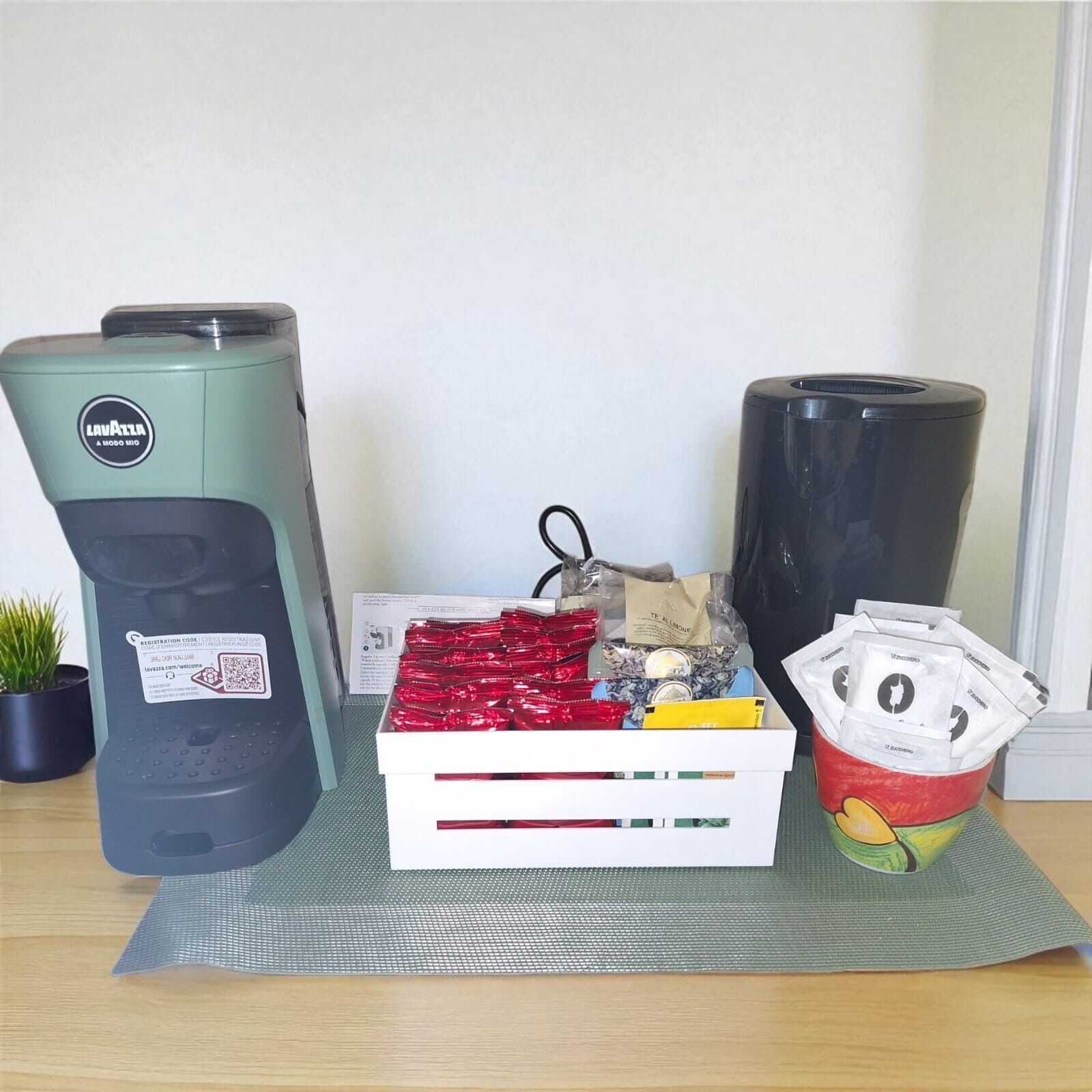 Eine Kaffeemaschine, eine Packung rote Teebeutel und ein roter Behälter mit gelbem Deckel auf einer Matte.
