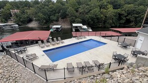 Una piscina con sillones y mesas, rodeada por una cerca, con un paisaje de colinas boscosas al fondo.
