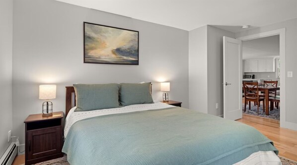 A bedroom with a bed, bedside lamps, a painting on the wall, and a view into the kitchen.