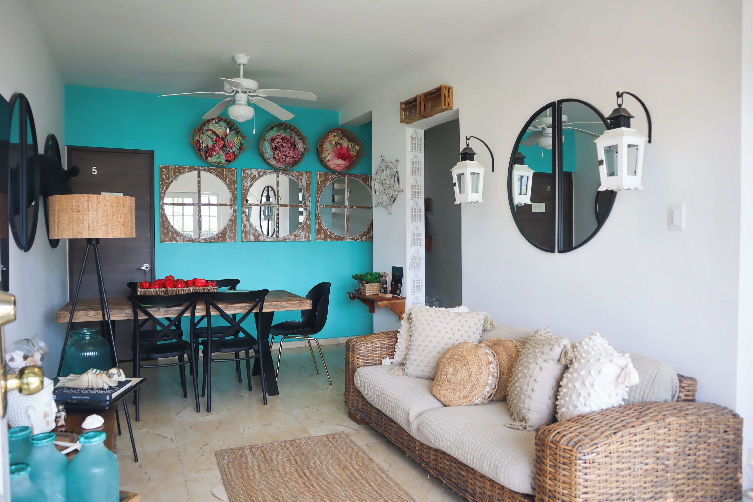 A room with a teal accent wall, a dining area with a wooden table and chairs, a wicker sofa with cushions, and a ceiling fan.