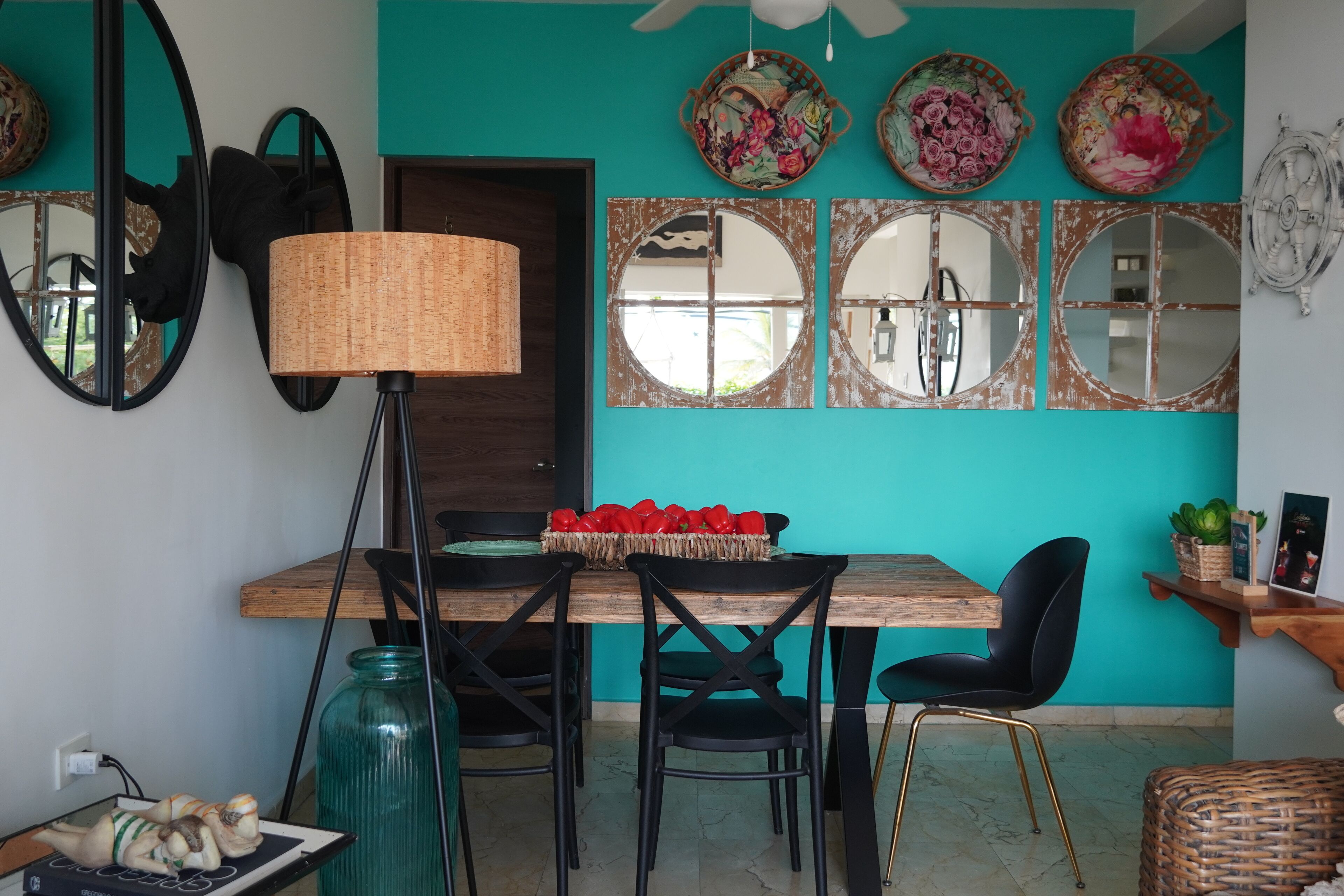 A dining area with a wooden table, black chairs, and a teal wall. There are decorative mirrors and a basket of red peppers on the table.