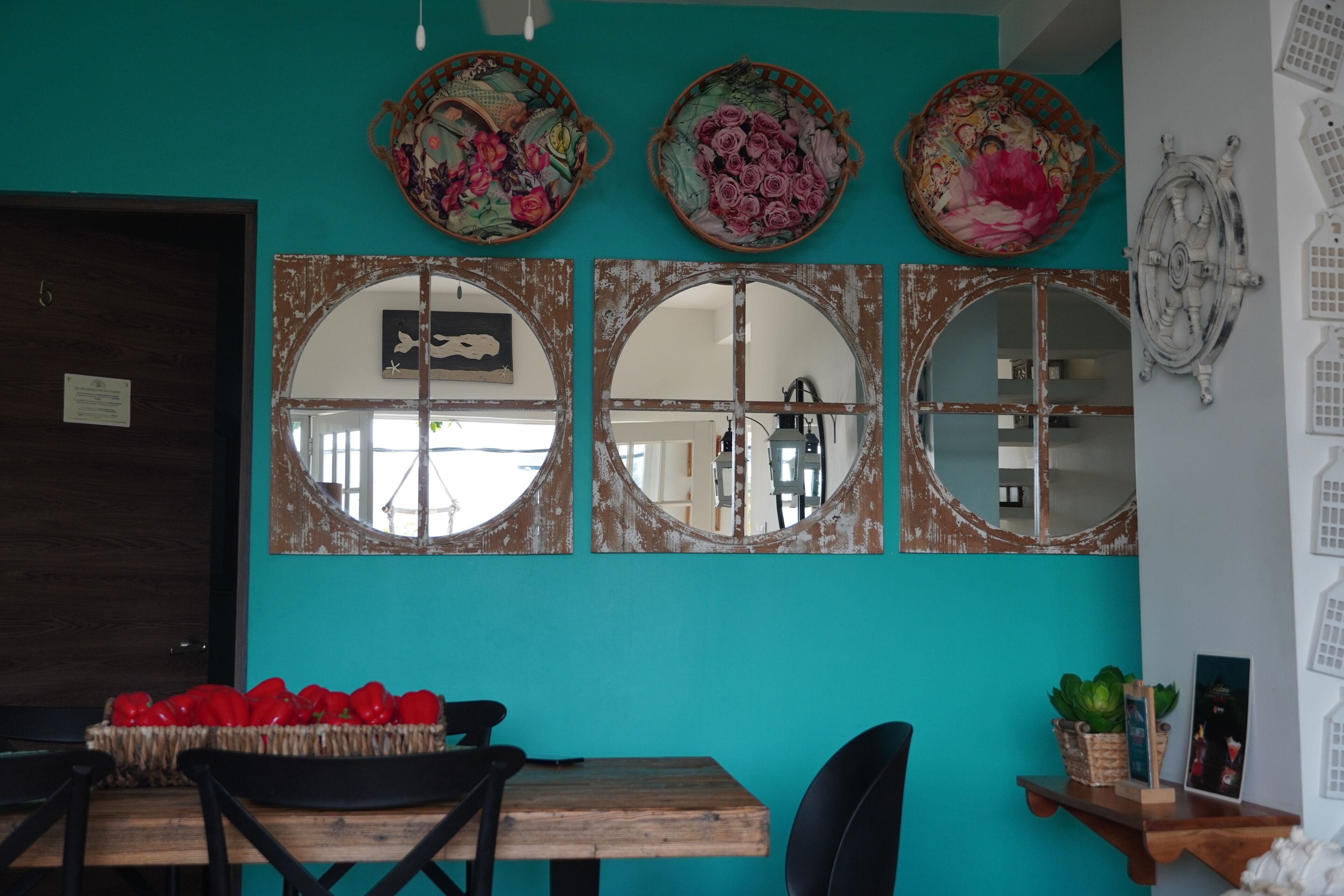 A dining area with a wooden table, chairs, and a basket of red peppers. There are three decorative mirrors on the wall, and a door with a sign on it.