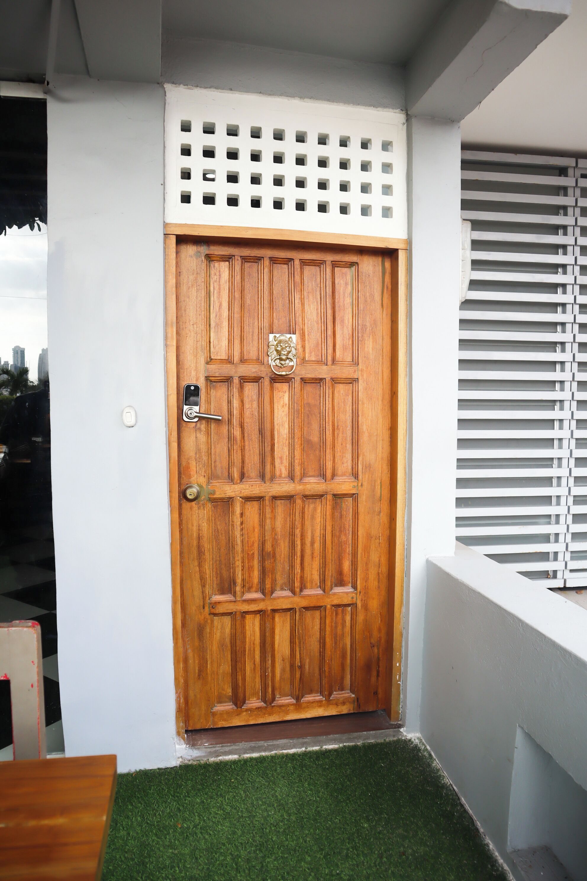 A wooden door with a keypad lock, a small window with horizontal blinds, and a white wall with a decorative panel above the door.