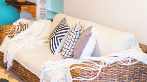 Un sofá de mimbre con una manta blanca y almohadas estampadas, un espejo redondo con dos faroles sujetos a la pared, y una mesita de madera con una planta.