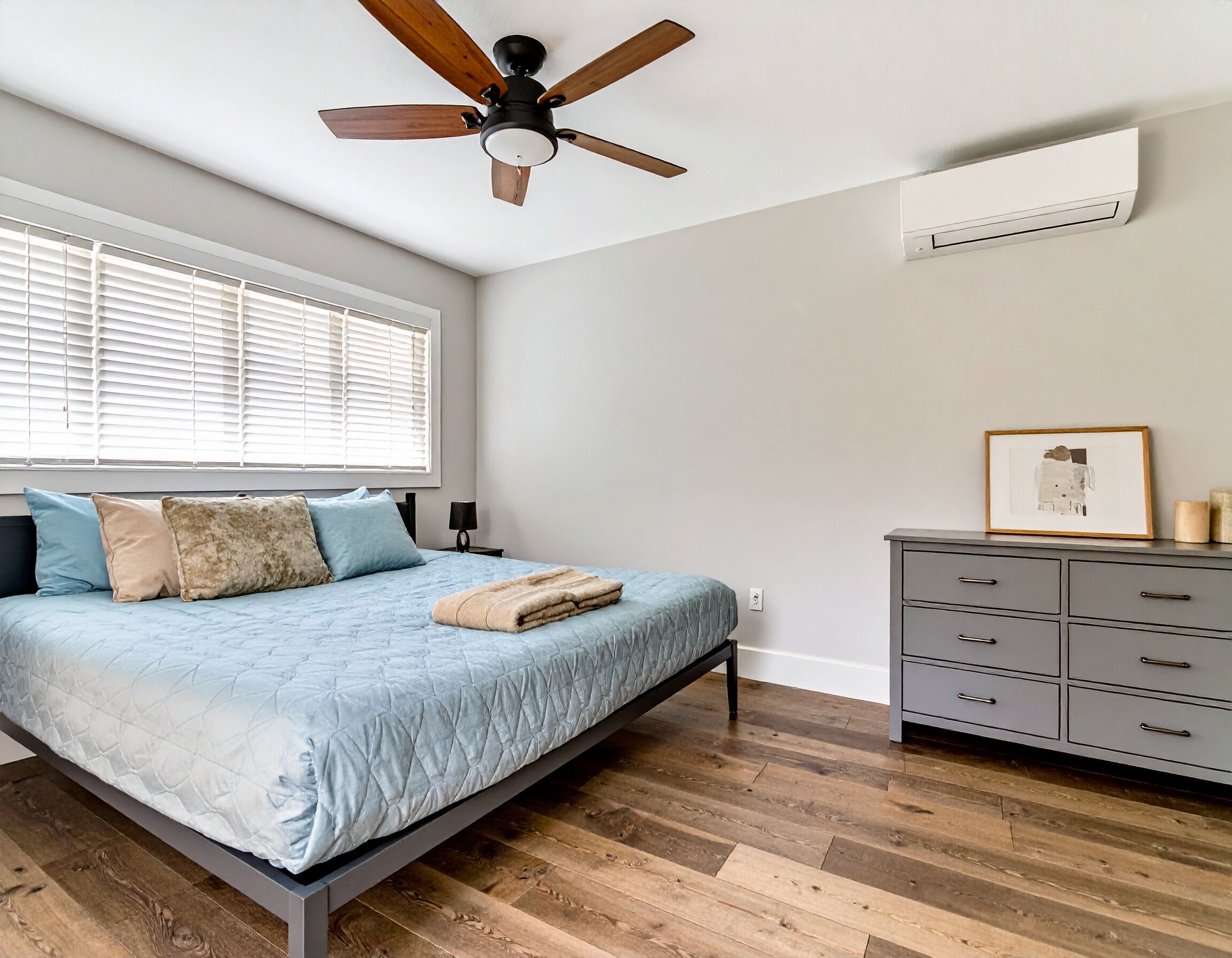 Une chambre à coucher comprenant un lit, un plancher en bois, un ventilateur de plafond, une fenêtre avec des stores, une commode et un appareil de climatisation.