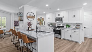 A modern kitchen with white cabinetry, a central island with bar stools, a microwave, oven, and a coffee bar. - Stay Local Fantastic 4 Bedroom Near the Rez Sleeps 16 (Brandon)
