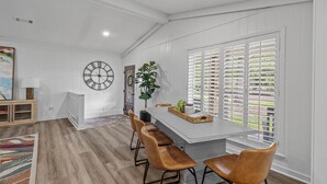 A dining area with a long table, four chairs, a clock, and a window with blinds. - Stay Local Fantastic 4 Bedroom Near the Rez Sleeps 16 (Brandon)
