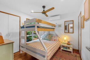A bunk bed room with a ceiling fan, a bedside table with a lamp, and a window with a view of greenery.