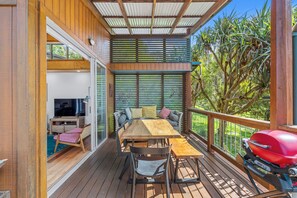 Een houten terras met een eettafel en stoelen, een barbecue en uitzicht op groen.