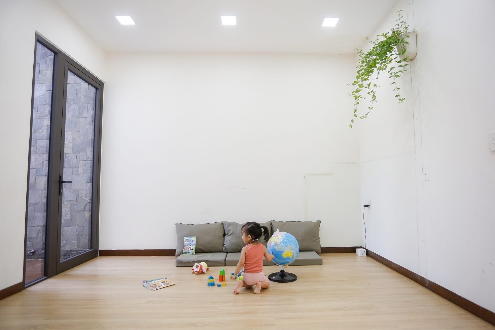 A child playing with toys on a wooden floor in a room with a grey sofa, a globe, and a hanging plant.