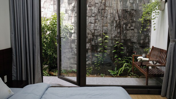 A bedroom with a bed, a bench, and a view of a stone wall and greenery outside. - Cozy 3BR Near Beach, Tranquil Fish Pond & Parking (Đà Nẵng)