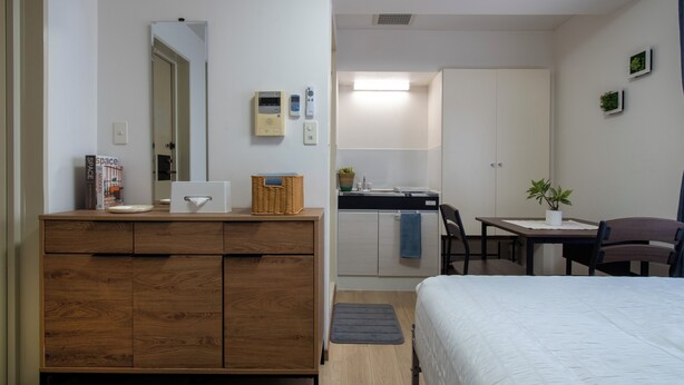 A compact hotel room with a wooden dresser, a bed, a desk with a chair, a small kitchenette, and a bathroom visible through an open door.