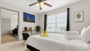 Une chambre avec un grand lit, un ventilateur de plafond, une fenêtre avec des stores, une porte donnant sur une autre pièce, et une petite table sur laquelle se trouvent une plante et un tableau.
