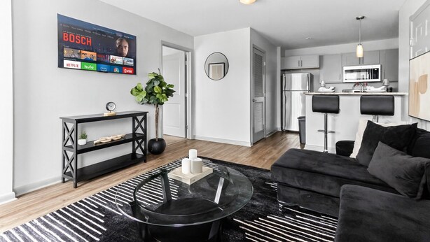 Un salon moderne avec un canapé noir, une table basse, un téléviseur fixé au mur qui diffuse différents services de streaming, une petite plante et un coin cuisine équipé d’un réfrigérateur et d’un four à micro-ondes.
