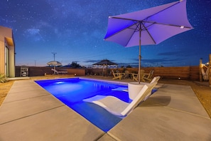 Une piscine dont le fond reflète un ciel étoilé, entourée de mobilier de jardin en bois.