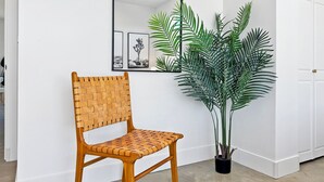 Une chaise en bois, dotée d’un dossier et d’une assise en fibres tressées, est placée à côté d’une plante en pot aux longues feuilles fines. Un miroir à cadre noir est accroché au mur, au-dessus de la chaise.