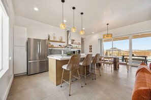 A modern kitchen with a stainless steel refrigerator, a bar area with wicker chairs, and a dining table with a view of the outdoors. - Desert Spa: Heated Pool · Hot Tub · Sauna · Cold Plunge · New Home - Joshua Tree (Joshua Tree)