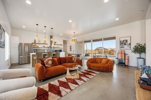 A spacious living area with a modern kitchen, a dining area, and a large window offering a view of the outdoors. - Desert Spa: Heated Pool · Hot Tub · Sauna · Cold Plunge · New Home - Joshua Tree (Joshua Tree)