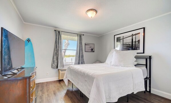 A bedroom with a bed, a dresser, a ceiling light, a window with curtains, and a framed picture on the wall.