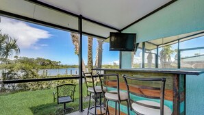 Un balcón con vista a un lago y palmeras, con una mesa y sillas, y un televisor montado.