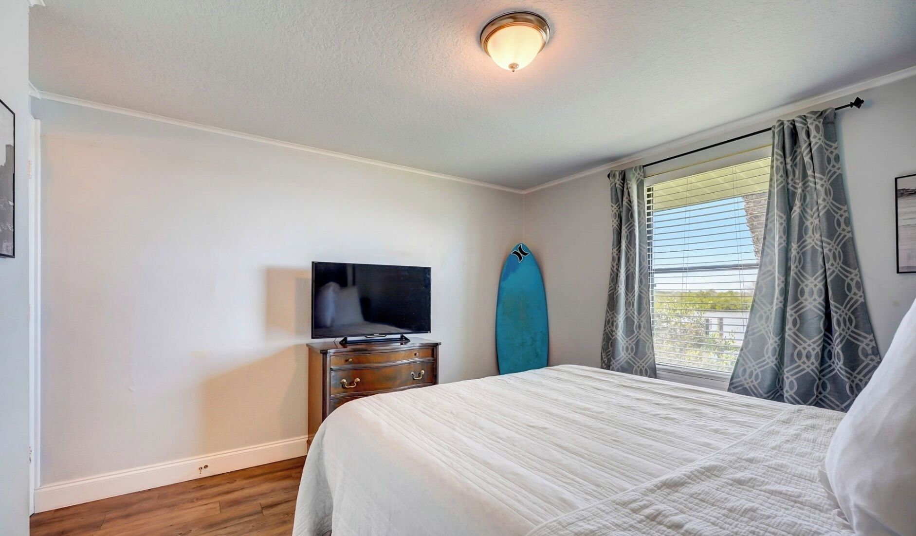A bedroom with a bed, a dresser, a television, and a surfboard against the wall.
