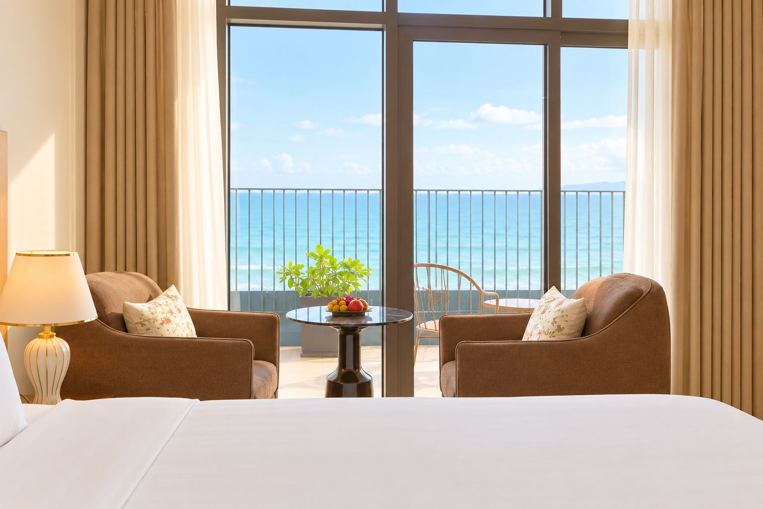 Ein Hotelzimmer mit einem großen Bett, zwei Sesseln, einem kleinen Tisch mit Obst und Meerblick durch raumhohe Fenster.