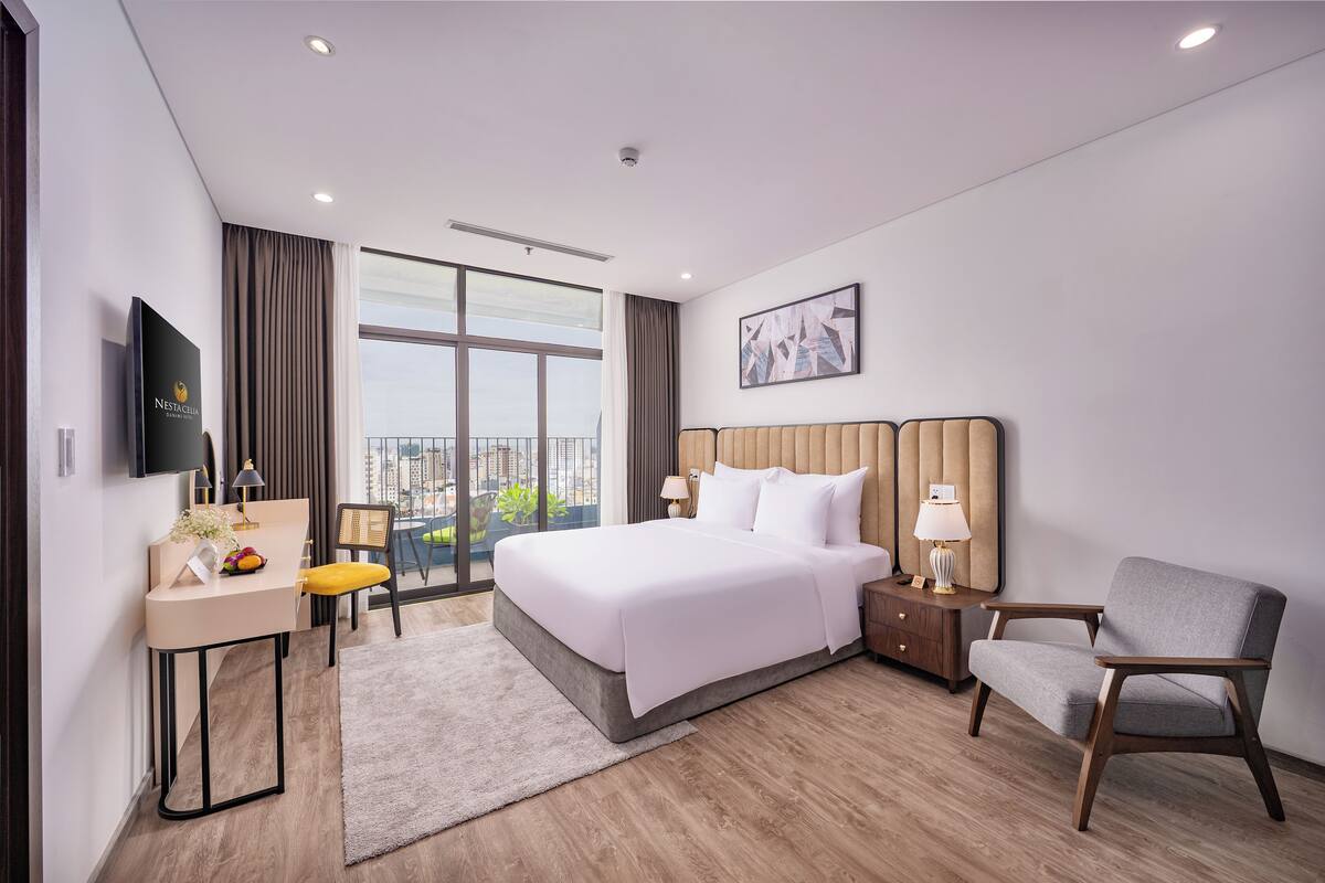Een moderne hotelkamer met een groot bed, een bureau met stoel, nachtkastje met lampen en uitzicht op de stad door grote ramen.