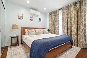 A bedroom with a wooden bed, a bedside table with a lamp, and a wall-mounted air conditioner. - Coastal Chic 2BR/1b / Long Beach (Long Beach)
