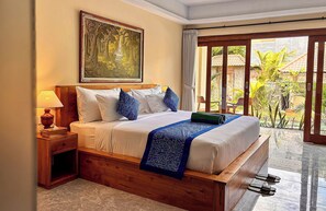 A bedroom with a wooden bed, white bedding, a blue patterned throw, a nightstand with a lamp, and a large window with a view of greenery. - Mimpi Luwih Ubud (Ubud)