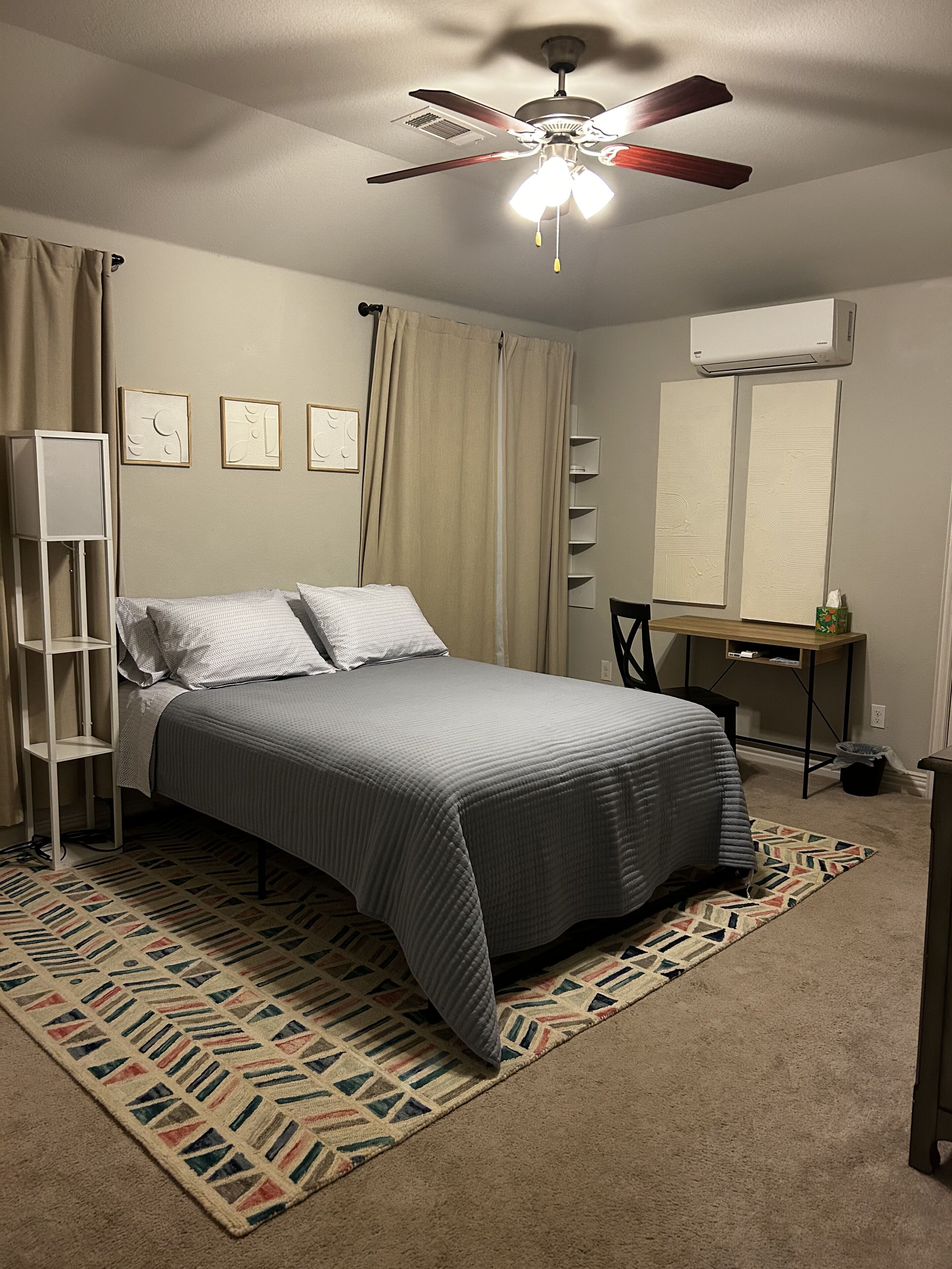 Een slaapkamer met een bed, een bureau, een stoel, een staande lamp, een plafondventilator en een kamerkleed met patroon.