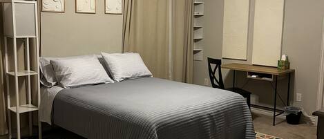 Een slaapkamer met een bed, een bureau, een stoel, een staande lamp, een plafondventilator en een kamerkleed met patroon.