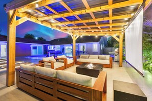 Un patio exterior con muebles de madera, una fogata y una piscina al fondo.
