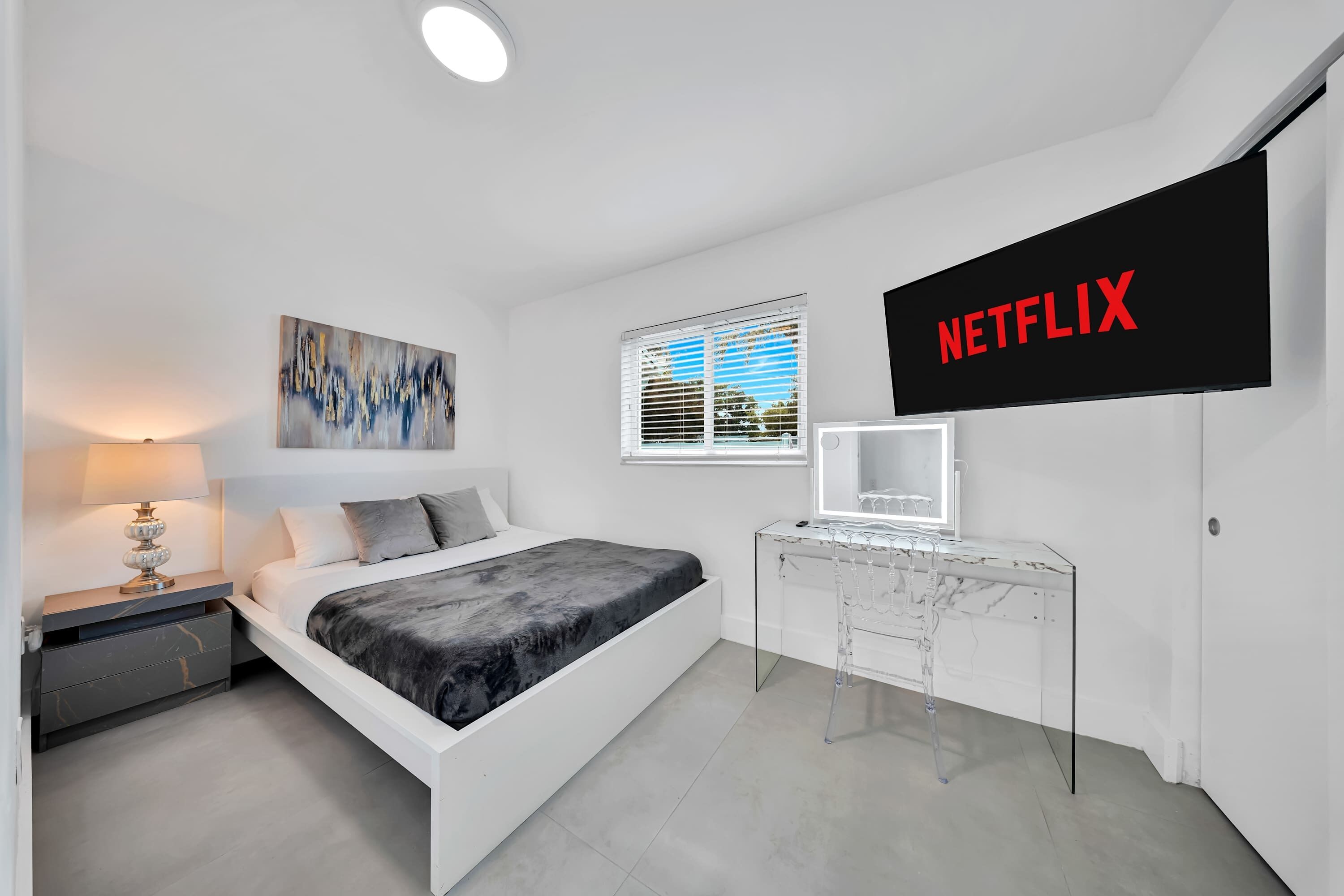 A modern bedroom with a bed, bedside table, lamp, artwork, window, and a wall-mounted TV labeled 'NETFLIX'.