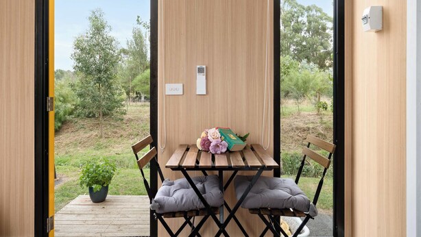 A wooden table with two cushioned chairs, a potted plant, and a box of flowers on a balcony with a view of greenery and trees.