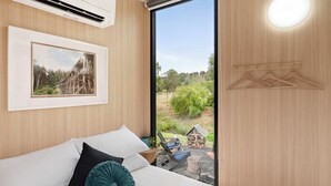 A bedroom with a bed, pillows, a framed picture, an air conditioner, a hanging light fixture, and a view of the outdoors.