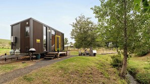 A modern, compact cabin with a large sliding door, a small porch area, and a gravel path leading to it.