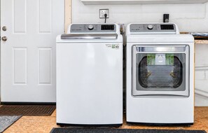 Two white front-loading washing machines in a laundry room with a white door and a power outlet on the wall. - Brandon Getaway | 4BR Home w/ Pool & Hot Tub (Brandon)