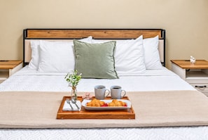 A neatly made bed with a tray of croissants and coffee cups, a vase with flowers, and a small figurine on a bedside table.