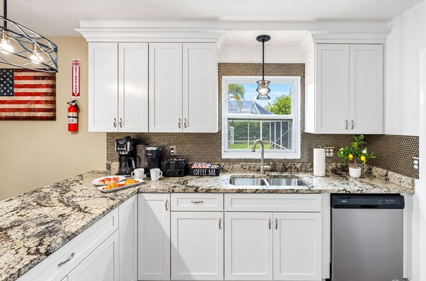 A kitchen with white cabinets, a granite countertop, a coffee bar, and a window with a view of a house and palm trees.