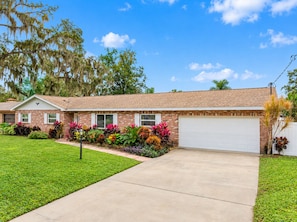 A single-story brick house with a white garage door, surrounded by well-maintained grass and colorful flowering plants.