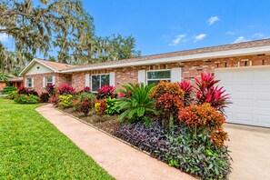 Una casa residencial con fachada de ladrillo, césped delantero y una variedad de plantas coloridas.