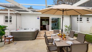 An outdoor patio area with a hot tub, a covered table with chairs, and a large umbrella. - Brandon Getaway | 4BR Home w/ Pool & Hot Tub (Brandon)